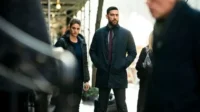 Pôster FBI:International:Agentes sérios,um homem barbudo e uma mulher de casaco preto,observam a rua urbana movimentada. Clima tenso e investigativo.