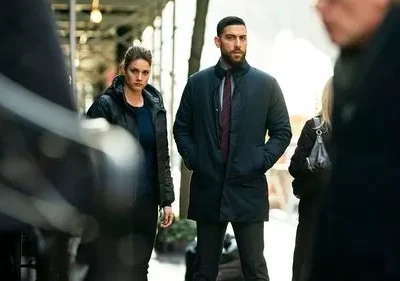 Pôster FBI: International: Agentes sérios, um homem barbudo e uma mulher de casaco preto, observam a rua urbana movimentada. Clima tenso e investigativo.