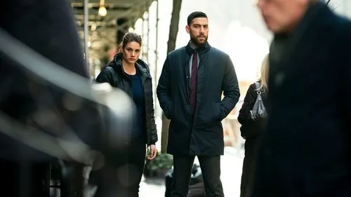 FBI – T01E16:Invisível 1 Pôster FBI:International:Agentes sérios,um homem barbudo e uma mulher de casaco preto,observam a rua urbana movimentada. Clima tenso e investigativo.