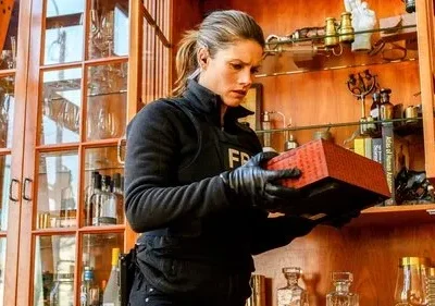 Uma agente do FBI, de luvas, examina uma caixa vermelha em um gabinete de madeira. Cena de investigação da série FBI.