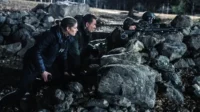 Agentes e atiradores em [Nome do Filme/Série] se abrigam atrás de uma parede de pedras,prontos para ação. Cena noturna tensa.
