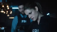 FBI:Dois agentes em coletes FBI em um ambiente escuro e tenso. A mulher em foco olha para baixo concentrada,homem ao fundo.