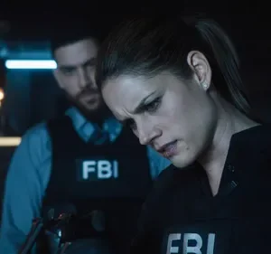 FBI: Dois agentes em coletes FBI em um ambiente escuro e tenso. A mulher em foco olha para baixo concentrada, homem ao fundo.