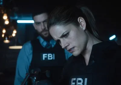 FBI: Dois agentes em coletes FBI em um ambiente escuro e tenso. A mulher em foco olha para baixo concentrada, homem ao fundo.