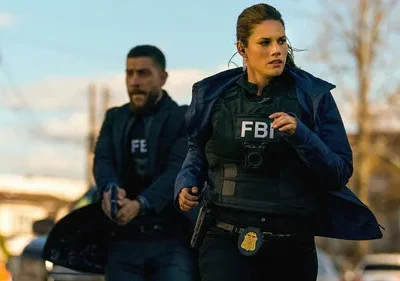 Agentes do FBI correm em uma rua urbana. A mulher no colete FBI e o homem com arma indicam uma cena de ação da série FBI.