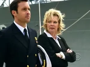 Hawaii Five-0: Steve McGarrett, em uniforme naval, e uma mulher loira séria, com um navio militar ao fundo.