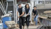 Aqui está uma opção de texto alternativo conciso:Em Hawaii Five-0,três agentes (dois homens,uma mulher) correm com coletes em área industrial,armas em punho. Ação tensa.
