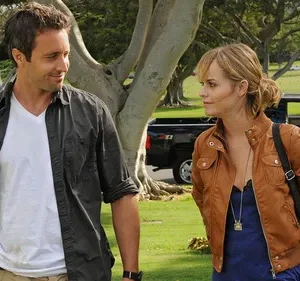 Em Hawaii Five-0, Alex O'Loughlin e Taryn Manning em um cemitério ensolarado. Ele sorri para ela, que tem olhar sério.