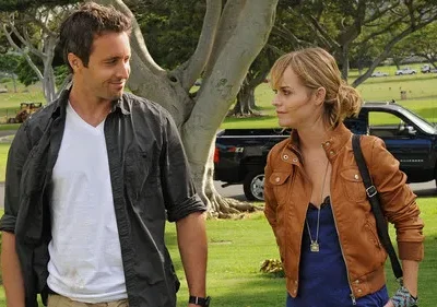 Em Hawaii Five-0, Alex O'Loughlin e Taryn Manning em um cemitério ensolarado. Ele sorri para ela, que tem olhar sério.