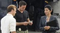 Hawaii Five-0:Steve e Danny analisam papéis,Kono em terno conversa. Equipe policial com viatura ao fundo,clima sério.