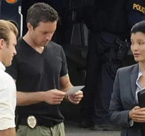 Hawaii Five-0: Steve e Danny analisam papéis, Kono em terno conversa. Equipe policial com viatura ao fundo, clima sério.