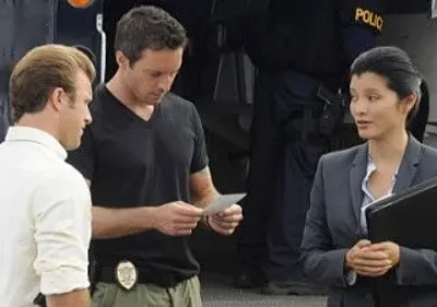 Hawaii Five-0: Steve e Danny analisam papéis, Kono em terno conversa. Equipe policial com viatura ao fundo, clima sério.