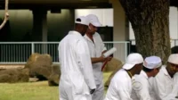 Aqui está uma opção concisa: Homens de macacão branco em Lost, alguns sentados, um em pé segurando um rolo de pintura, em um acampamento. Clima de trabalho ou cativeiro.