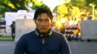 Daniel Dae Kim (Chin Ho Kelly) em Hawaii Five-0,com olhar sério,camisa azul e fone de pescoço,em cenário externo de ação.