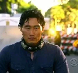 Daniel Dae Kim (Chin Ho Kelly) em Hawaii Five-0, com olhar sério, camisa azul e fone de pescoço, em cenário externo de ação.