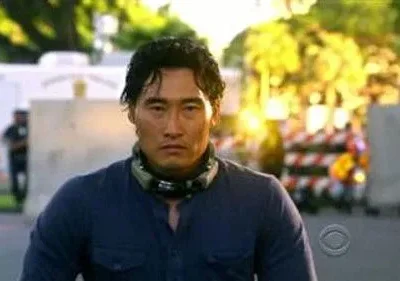 Daniel Dae Kim (Chin Ho Kelly) em Hawaii Five-0, com olhar sério, camisa azul e fone de pescoço, em cenário externo de ação.