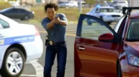 Chin Ho Kelly (Hawaii Five-0) mira uma arma,em cenário urbano com carros da polícia. Clima de ação e perigo.