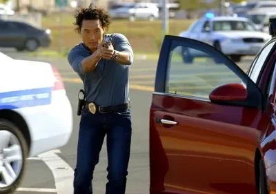 Chin Ho Kelly (Hawaii Five-0) mira uma arma, em cenário urbano com carros da polícia. Clima de ação e perigo.
