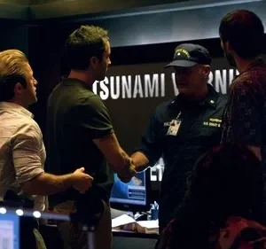 Hawaii Five-0: Homens numa sala de controle. Um oficial da Guarda Costeira aperta a mão de um agente, com TSUNAMI WARNING na tela. Tensão e urgência.