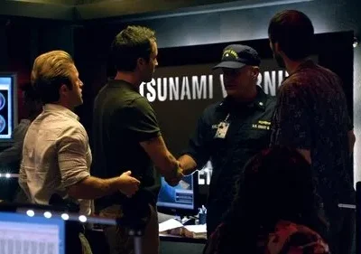 Hawaii Five-0: Homens numa sala de controle. Um oficial da Guarda Costeira aperta a mão de um agente, com TSUNAMI WARNING na tela. Tensão e urgência.