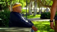Havaí Cinco-0 – T01E17:Powa Maka Moana (Piratas) 8 Hurley de Lost,um homem corpulento com bandana amarela,sentado num banco em um parque tropical ensolarado,olhando à direita. Uma cena calma.