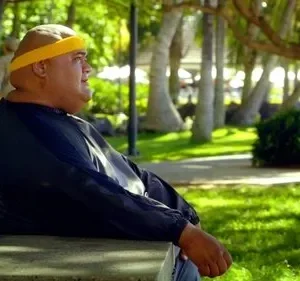Hurley de Lost, um homem corpulento com bandana amarela, sentado num banco em um parque tropical ensolarado, olhando à direita. Uma cena calma.