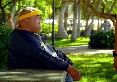 Hurley de Lost, um homem corpulento com bandana amarela, sentado num banco em um parque tropical ensolarado, olhando à direita. Uma cena calma.