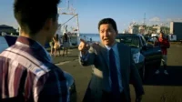 Cena de [Nome do Filme/Série]:Homem de terno asiático aponta firmemente para jovem de camisa xadrez em porto ensolarado. Barcos e SUV ao fundo.