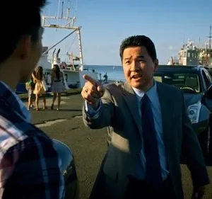 Cena de [Nome do Filme/Série]: Homem de terno asiático aponta firmemente para jovem de camisa xadrez em porto ensolarado. Barcos e SUV ao fundo.