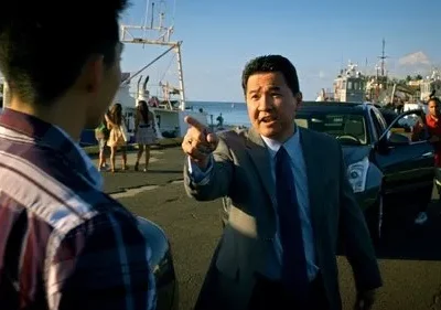 Cena de [Nome do Filme/Série]: Homem de terno asiático aponta firmemente para jovem de camisa xadrez em porto ensolarado. Barcos e SUV ao fundo.