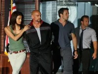 Havaí Cinco-0 – T01E19:Ne Me'e Laua Na Paio (Heróis e Vilões) 6 Cena de Hawaii Five-0:Kono escolta um suspeito enquanto McGarrett e Danno observam. Ação policial tensa em ambiente interno com bandeira.