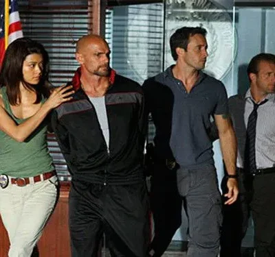 Cena de Hawaii Five-0: Kono escolta um suspeito enquanto McGarrett e Danno observam. Ação policial tensa em ambiente interno com bandeira.