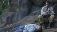 Havaí Cinco-0 – T01E20:Ma Ke Kahakai (Praia) 5 Hawaii Five-0:Steve McGarrett agachado sobre um homem inconsciente em terreno rochoso,segurando um rádio. Cena de tensão ao ar livre.