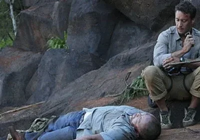 Hawaii Five-0: Steve McGarrett agachado sobre um homem inconsciente em terreno rochoso, segurando um rádio. Cena de tensão ao ar livre.