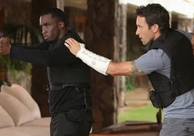 Hawaii Five-0: Diddy, armado e de colete tático, aponta pistola. McGarrett, com braço engessado, o guia. Cena de ação tensa.