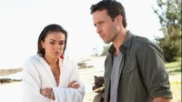 Havaí Cinco-0 – T01E22:Ho'ohuli Na'au (Perto do Coração) 3 Em Hawaii Five-0,um homem e uma mulher de roupão,ambos com expressão séria,num cenário de praia ensolarada. Clima tenso.