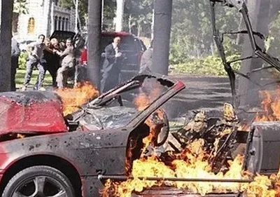 Pôster de [Nome do Filme/Série]: Carro vermelho em chamas intensas no primeiro plano. Pessoas correndo e em pânico ao fundo. Cena de ação e caos.