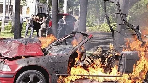Pôster de [Nome do Filme/Série]:Carro vermelho em chamas intensas no primeiro plano. Pessoas correndo e em pânico ao fundo. Cena de ação e caos.